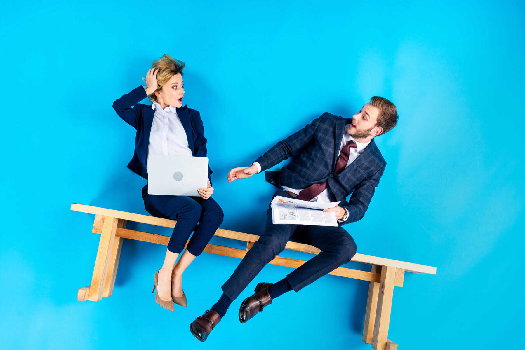 Un homme et une femme en tenue professionnelle sont assis sur un banc instable sur un fond bleu, exprimant la surprise. L'homme tient un journal et la femme un ordinateur portable, illustrant le concept de la gestion du risque de change et l'incertitude financière.
